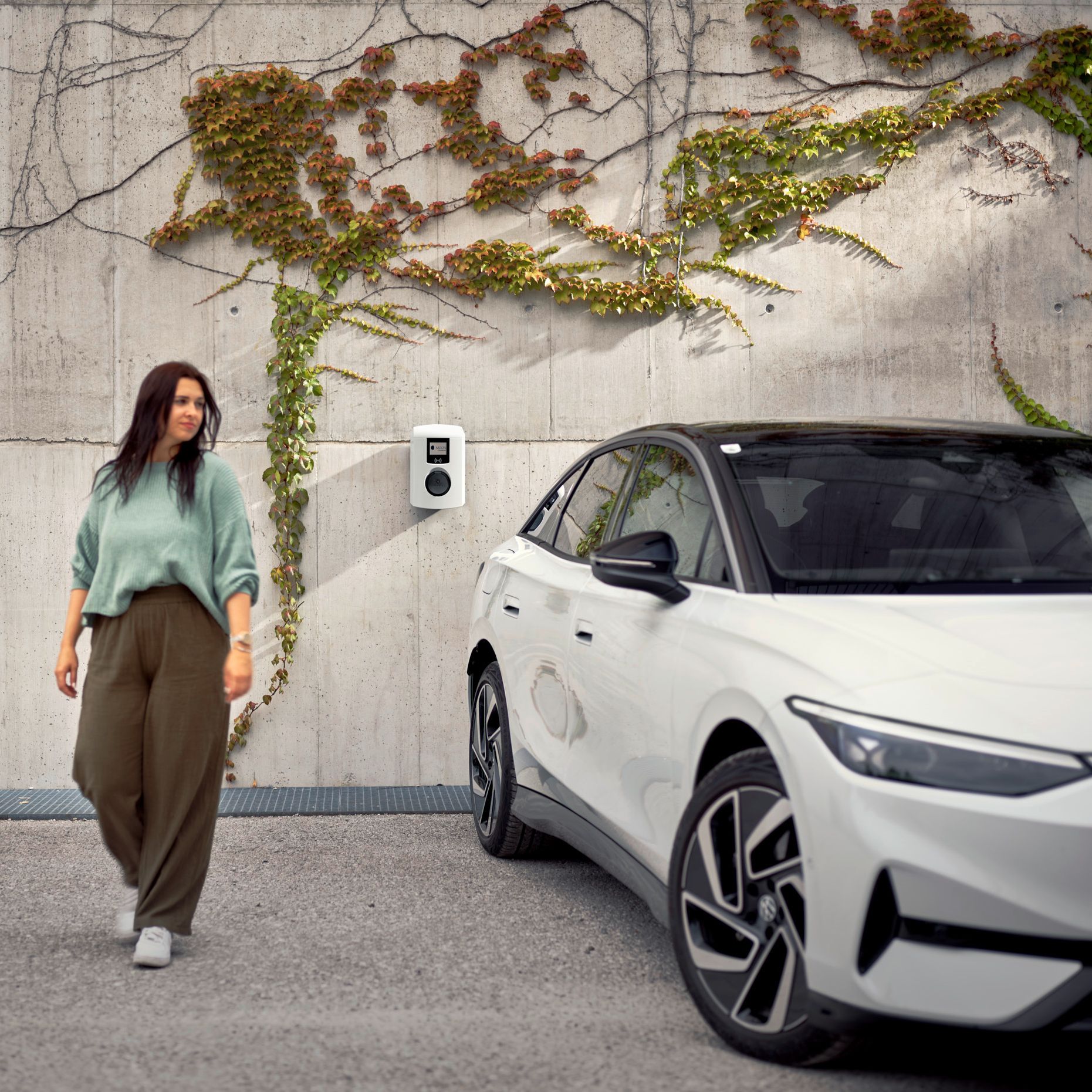 Woman walking next to an electric car in front of a wallbox towards the camera