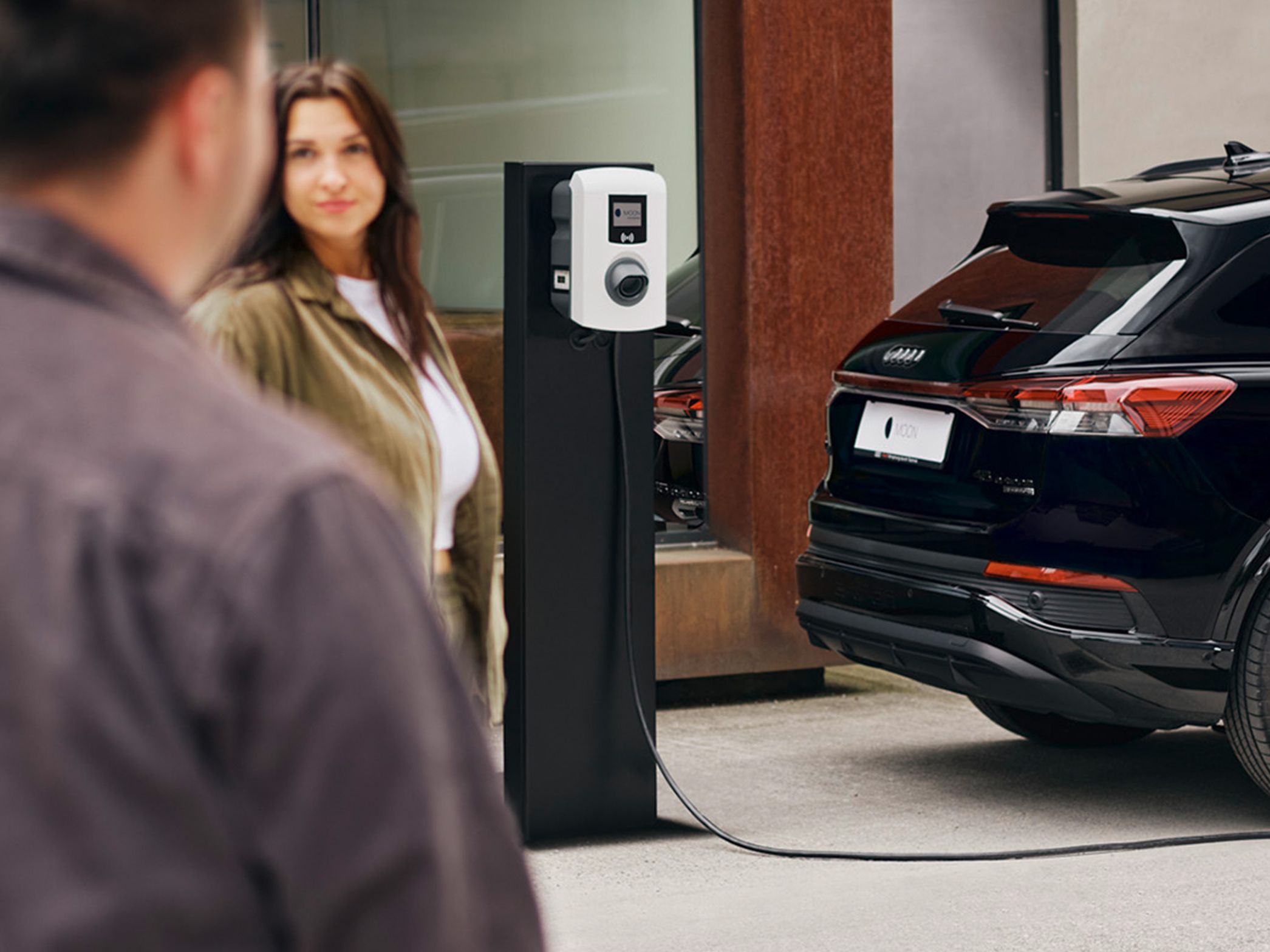 A car charges at a charging station with two people in the foreground