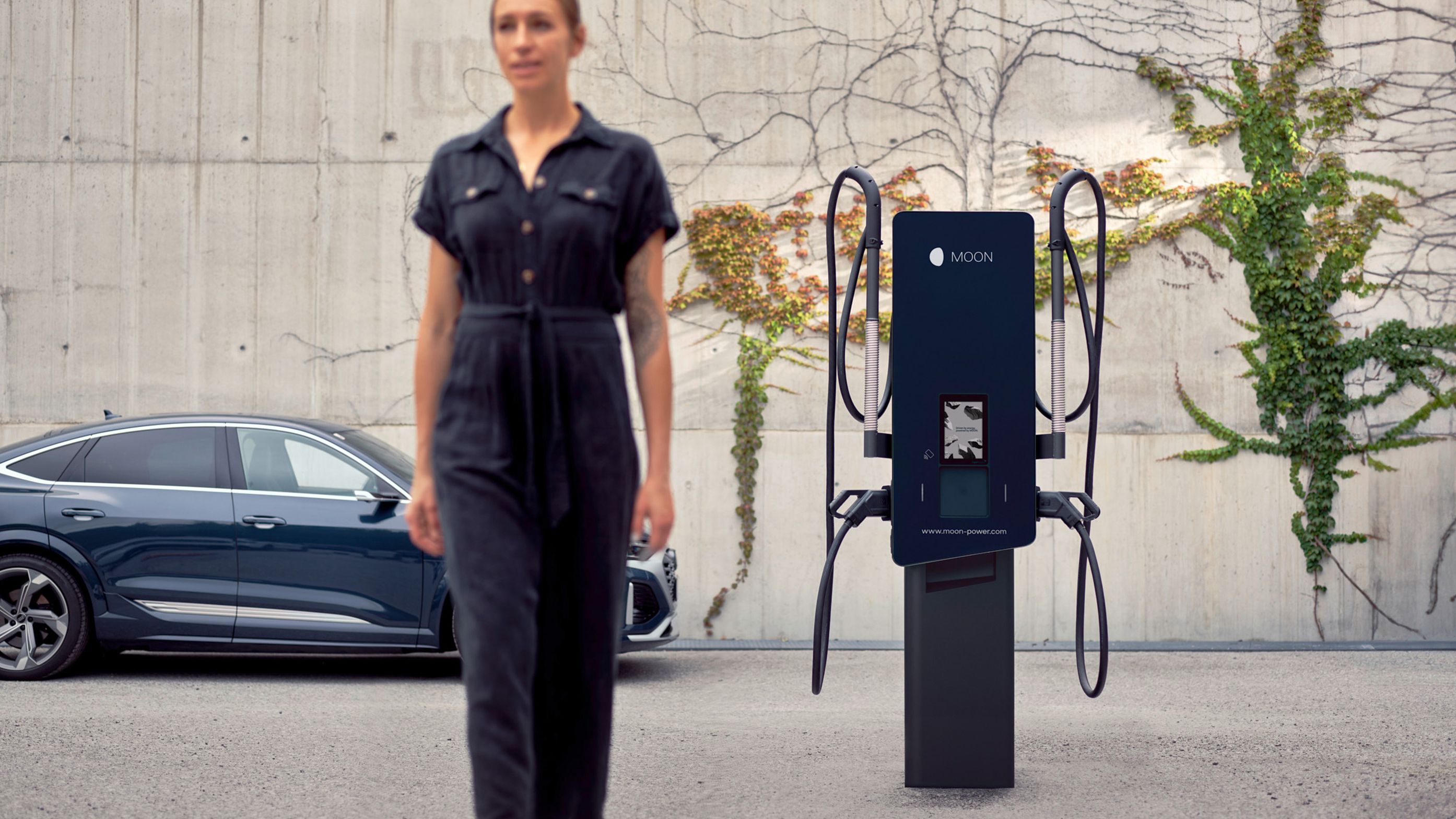 Woman walks in front of a wallbox with an electric car in the background