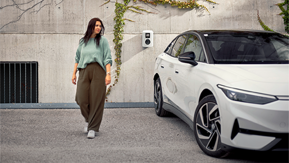 Woman walking next to an electric car in front of a wallbox towards the camera