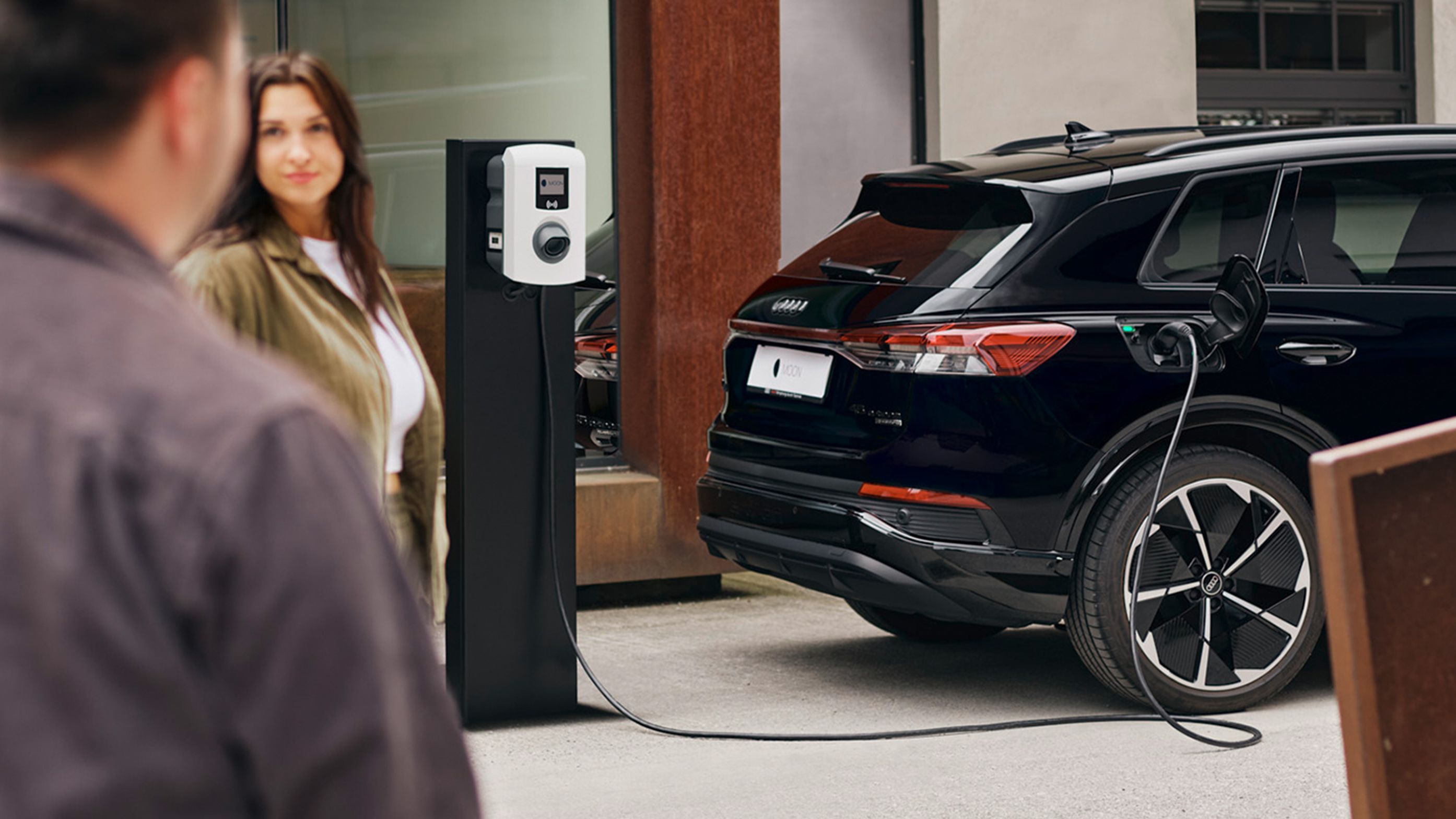 An electric car charges at a wallbox with two people in the foreground