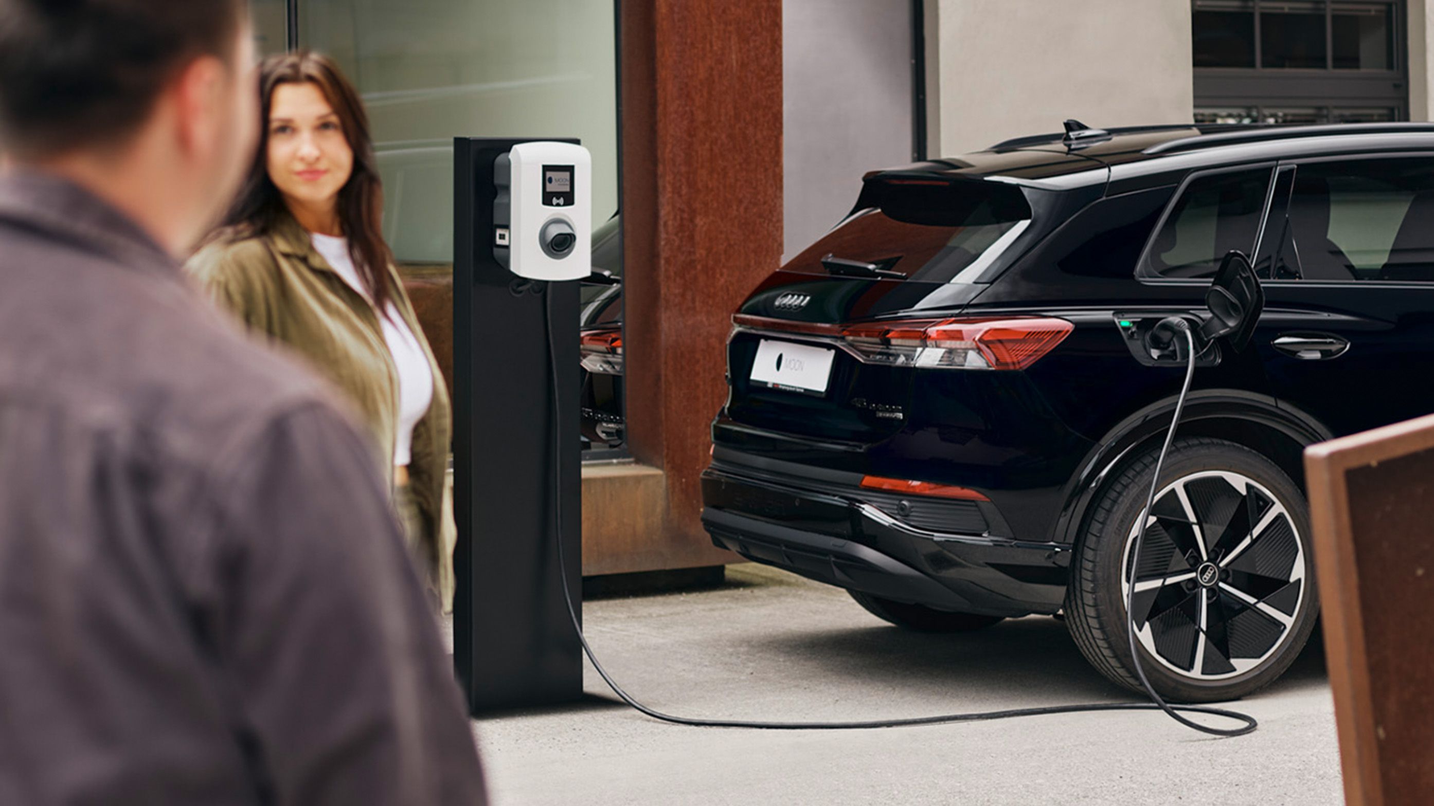 A car charges at a charging station with two people in the foreground