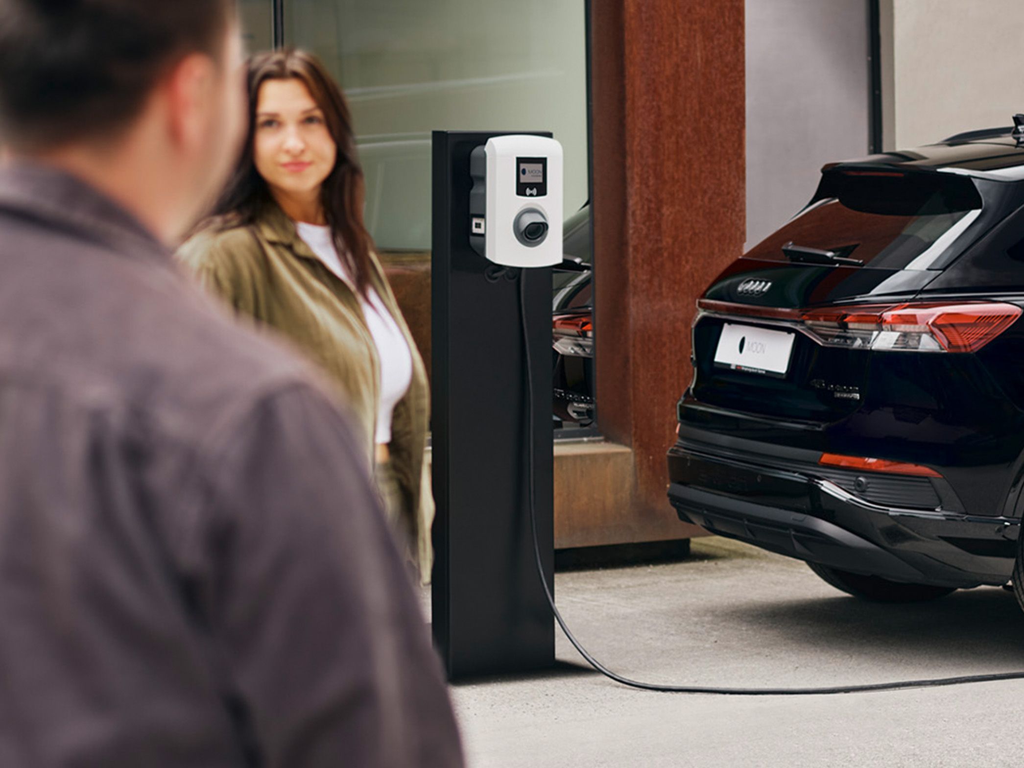 A car charges at a charging station with two people in the foreground
