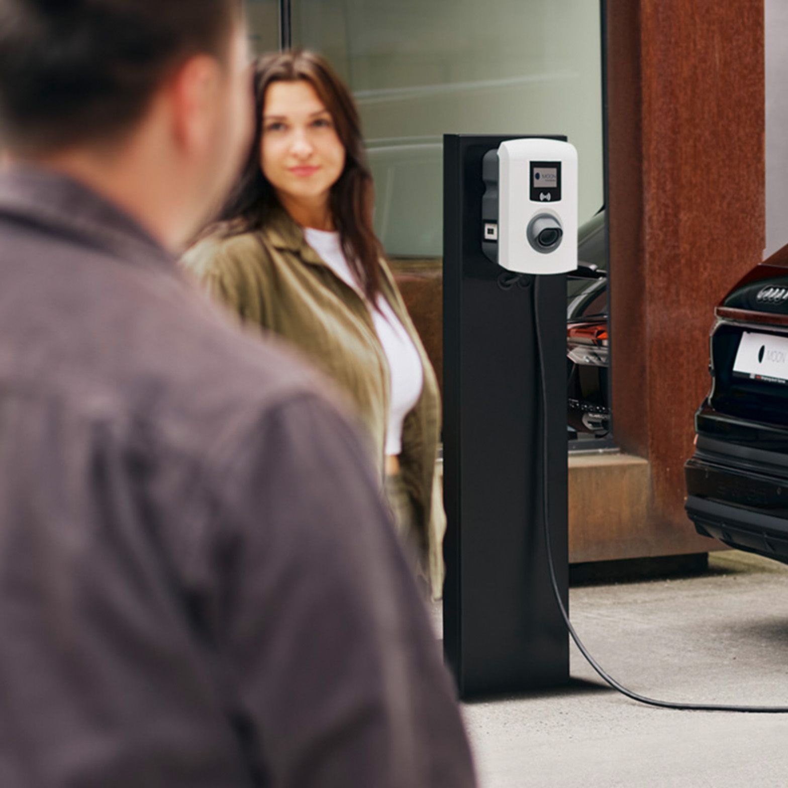 A car charges at a charging station with two people in the foreground
