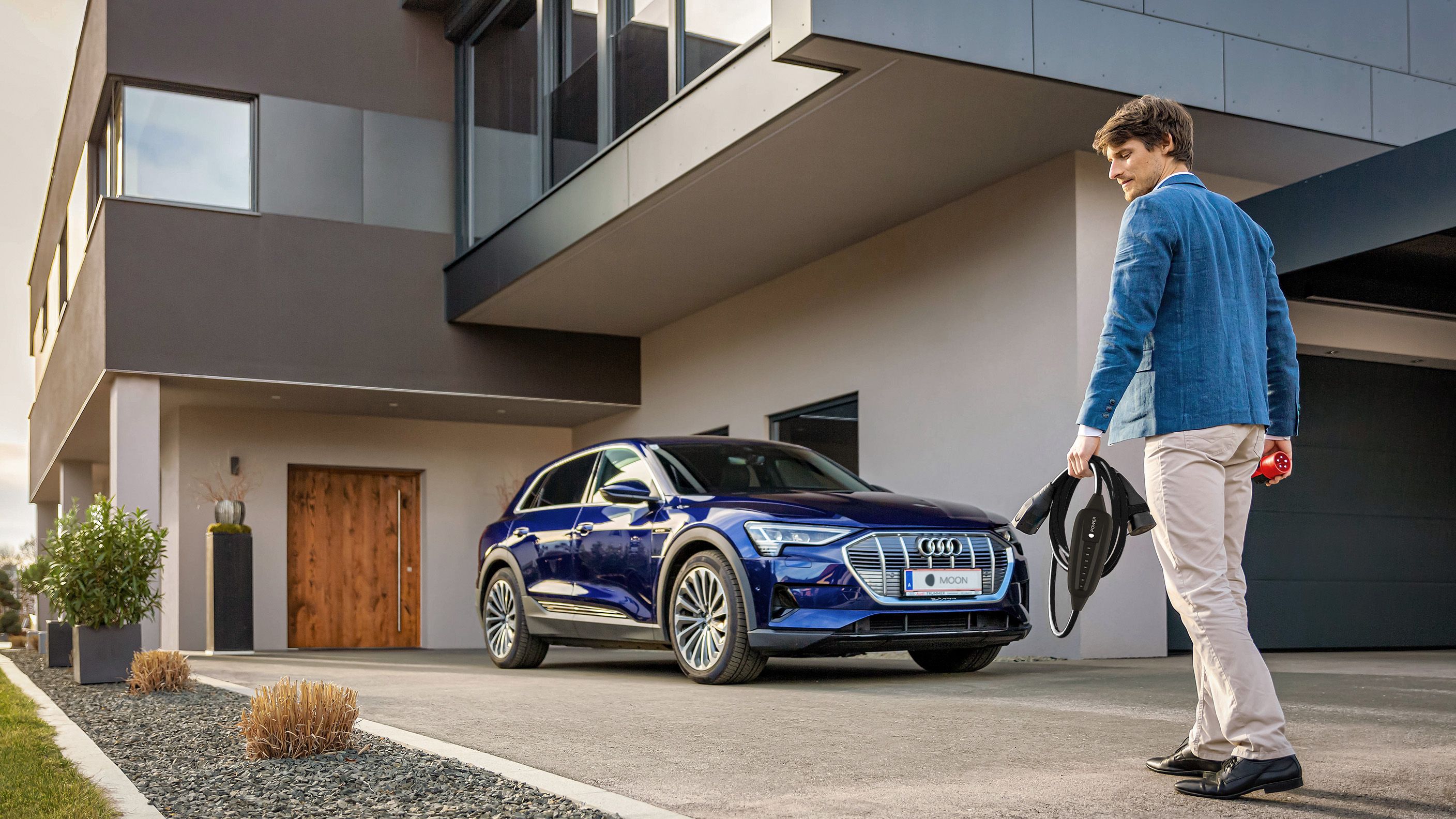 Man walks towards an electric car near a house with a cable in his hand