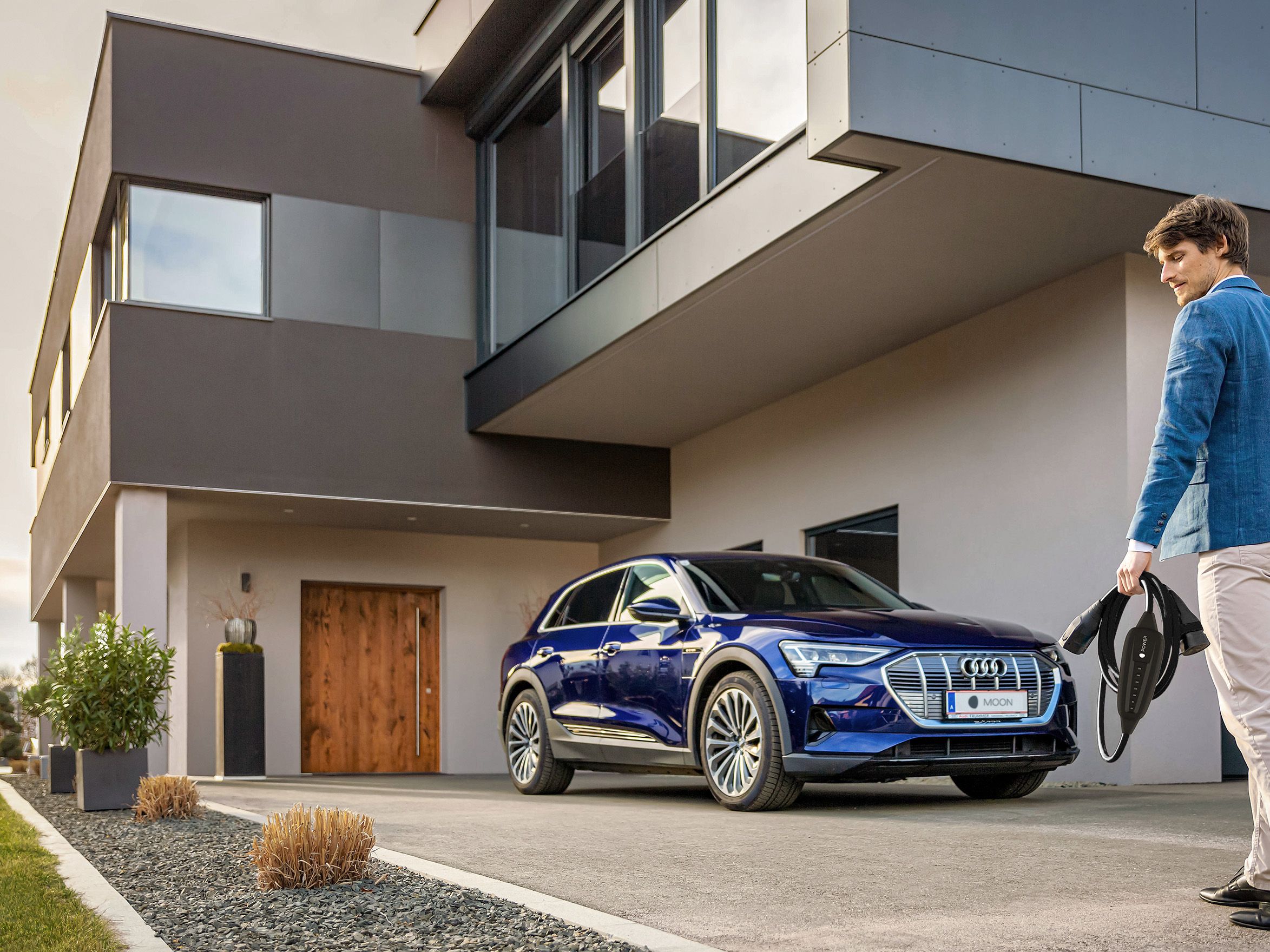 Man walks towards an electric car near a house with a cable in his hand