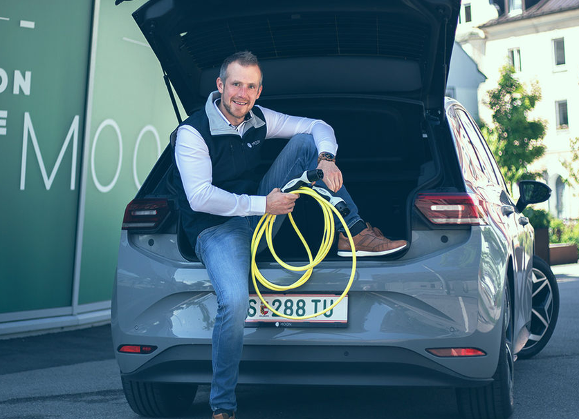 Support and Maintenance. Man sitting on the trunk of an e-car holding a charging cable."